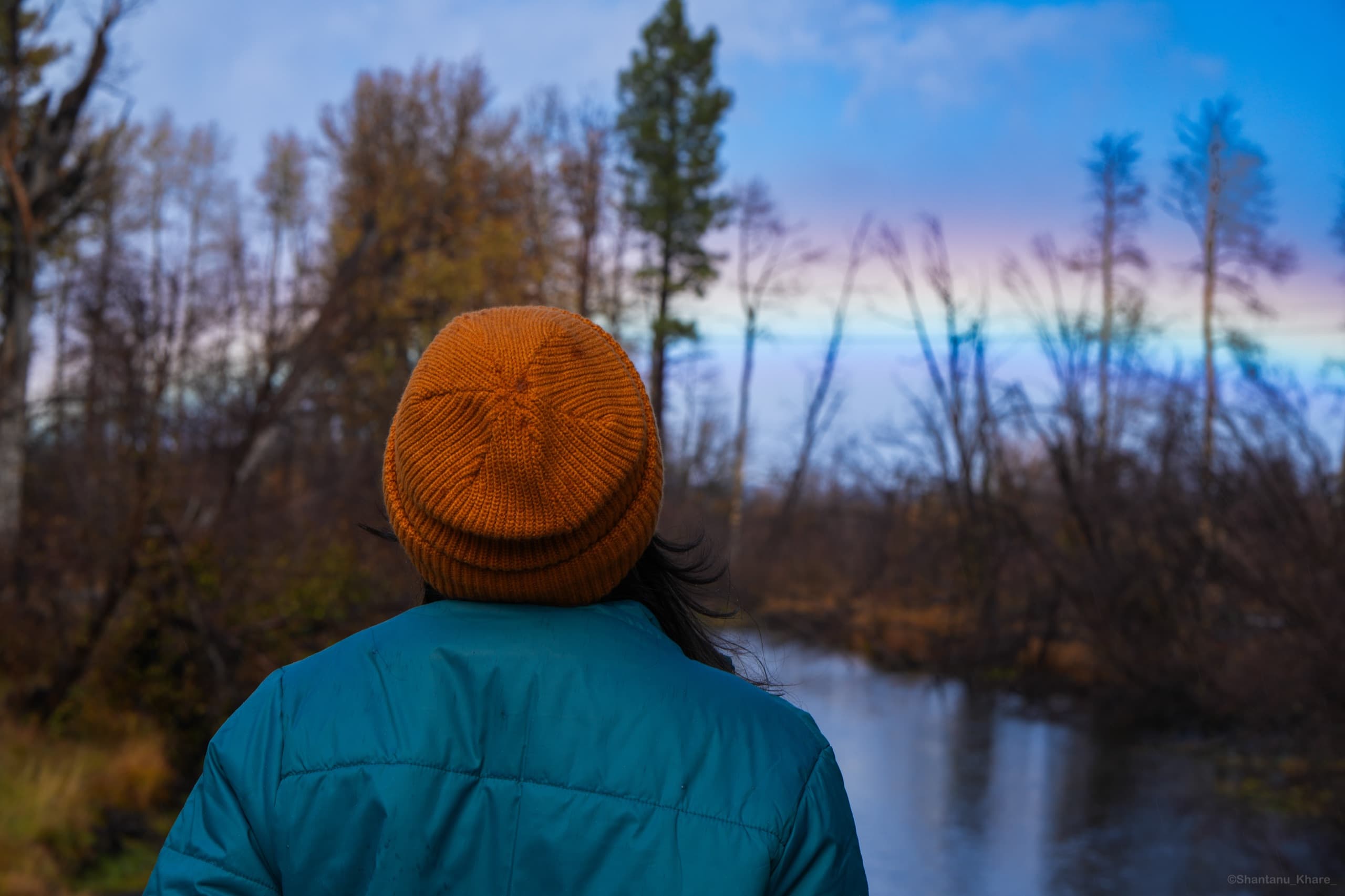 Travel photography — a person in an orange beanie gazes across a still river through autumn trees at dusk.