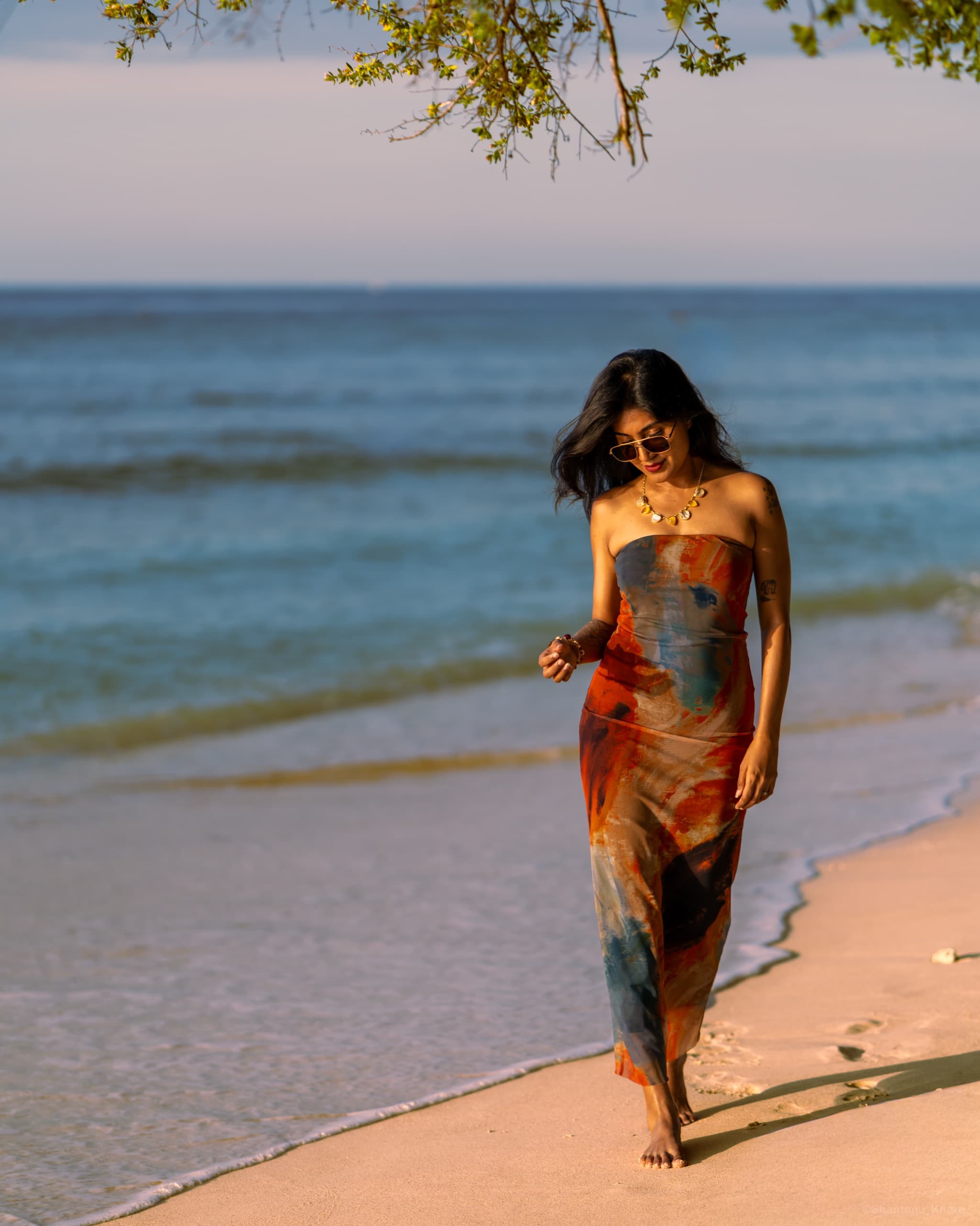 Portrait photography — a woman in a flowing printed dress walks a sunlit tropical beach.