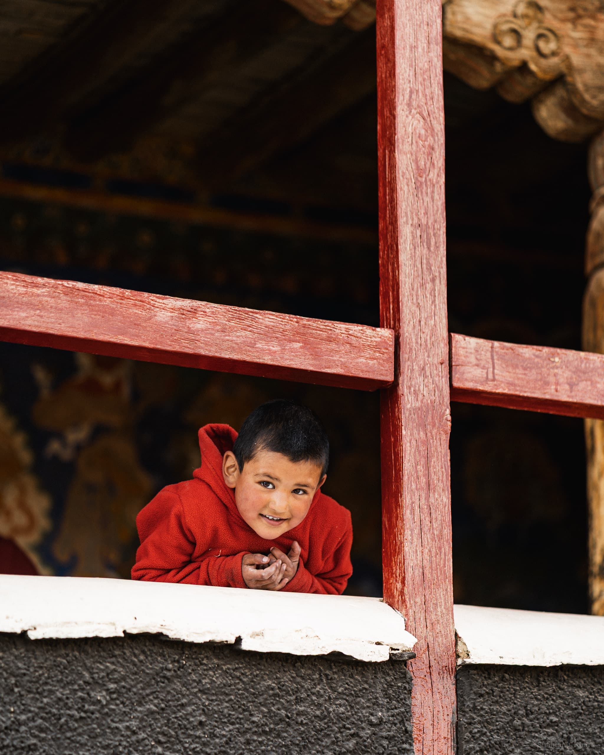 Documentary portrait photography — a young monk smiles from a painted monastery window in the Himalayas, Ladakh.