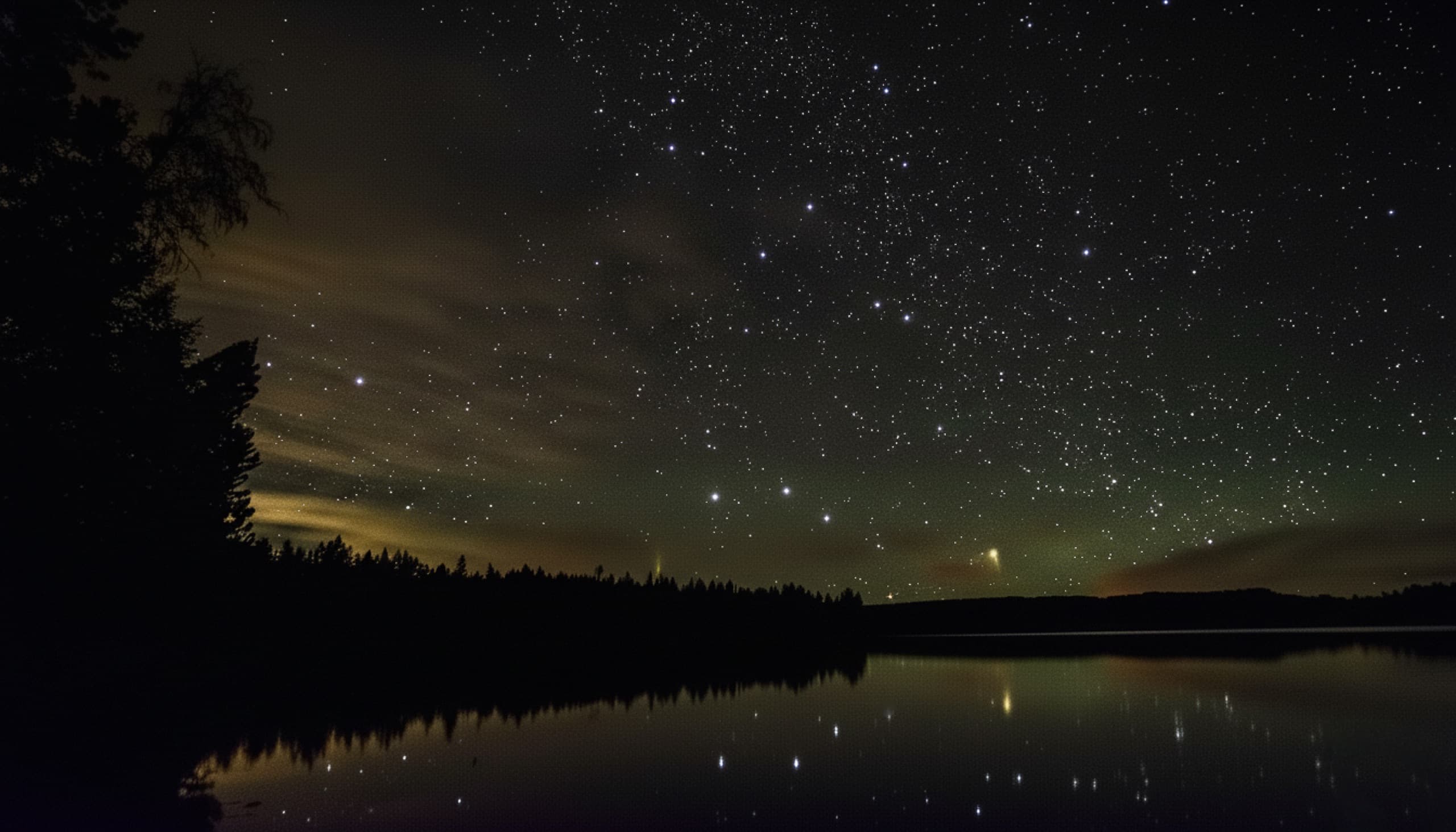 Astrophotography — a star-filled night sky reflected in a still lake ringed by dark forest.