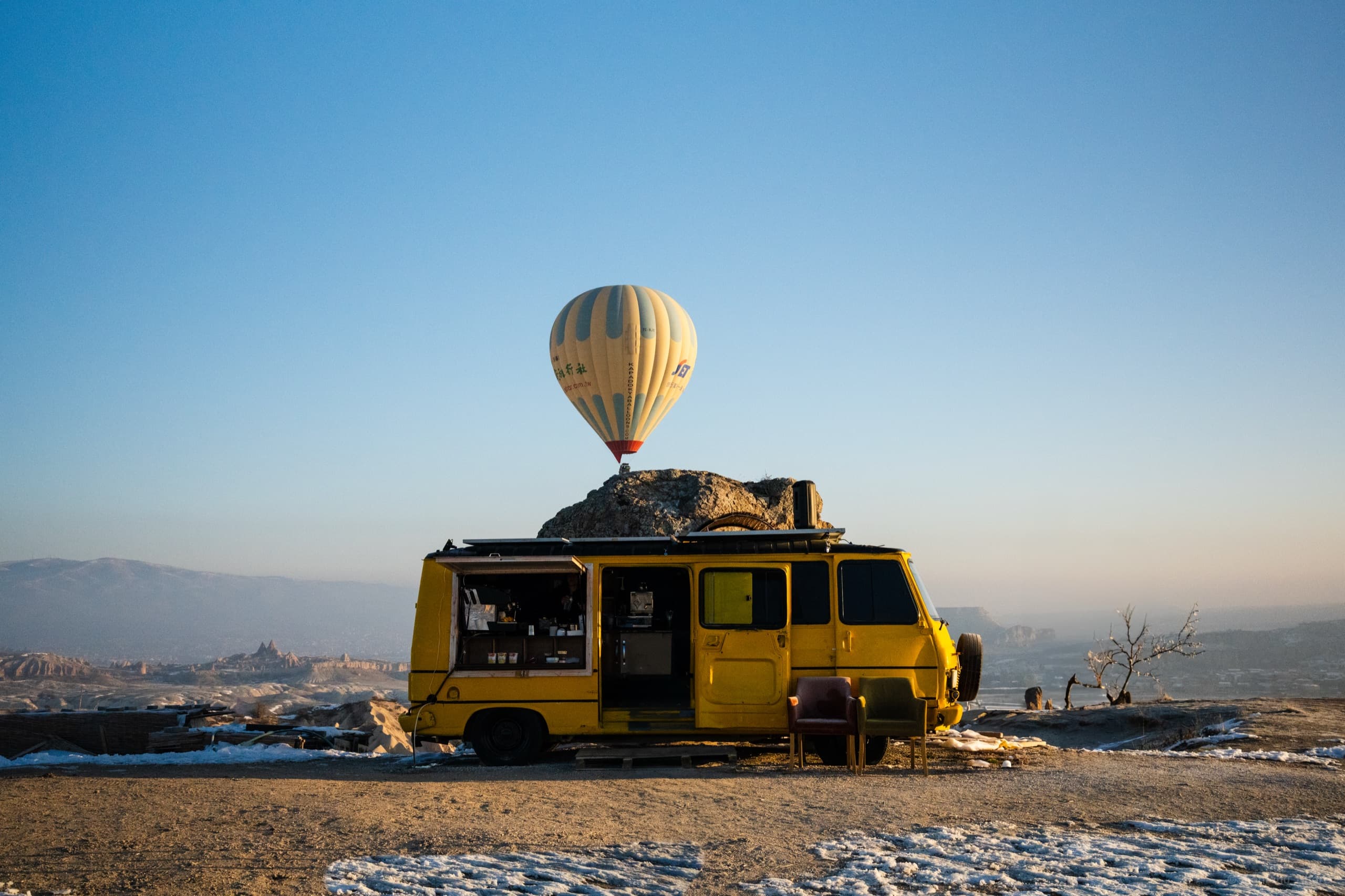 Travel photography — a hot-air balloon rising behind a yellow vintage van on a rocky overlook, Cappadocia.