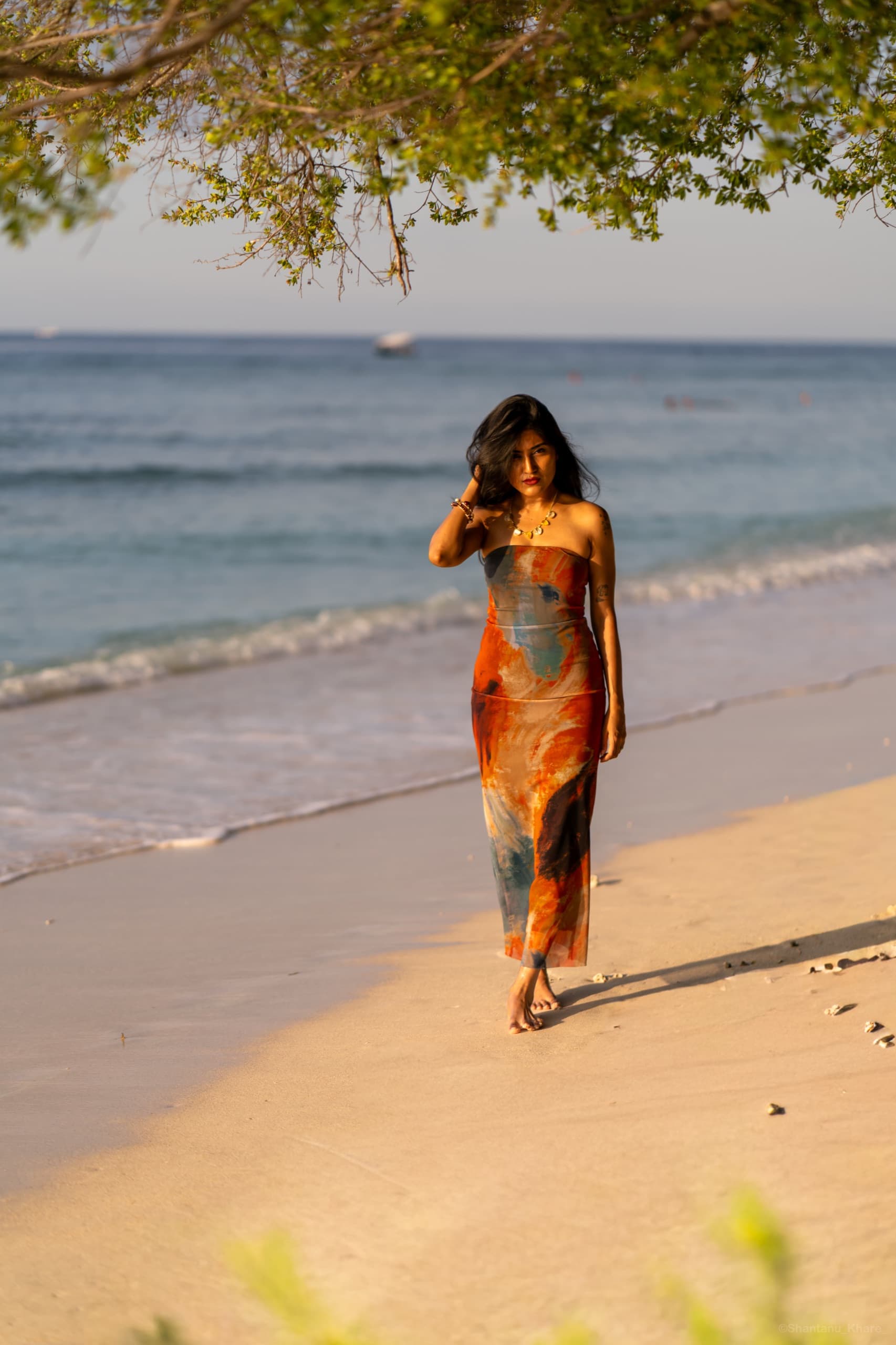 Portrait photography — a woman in a flowing printed dress walks the water's edge under a leaning tree.